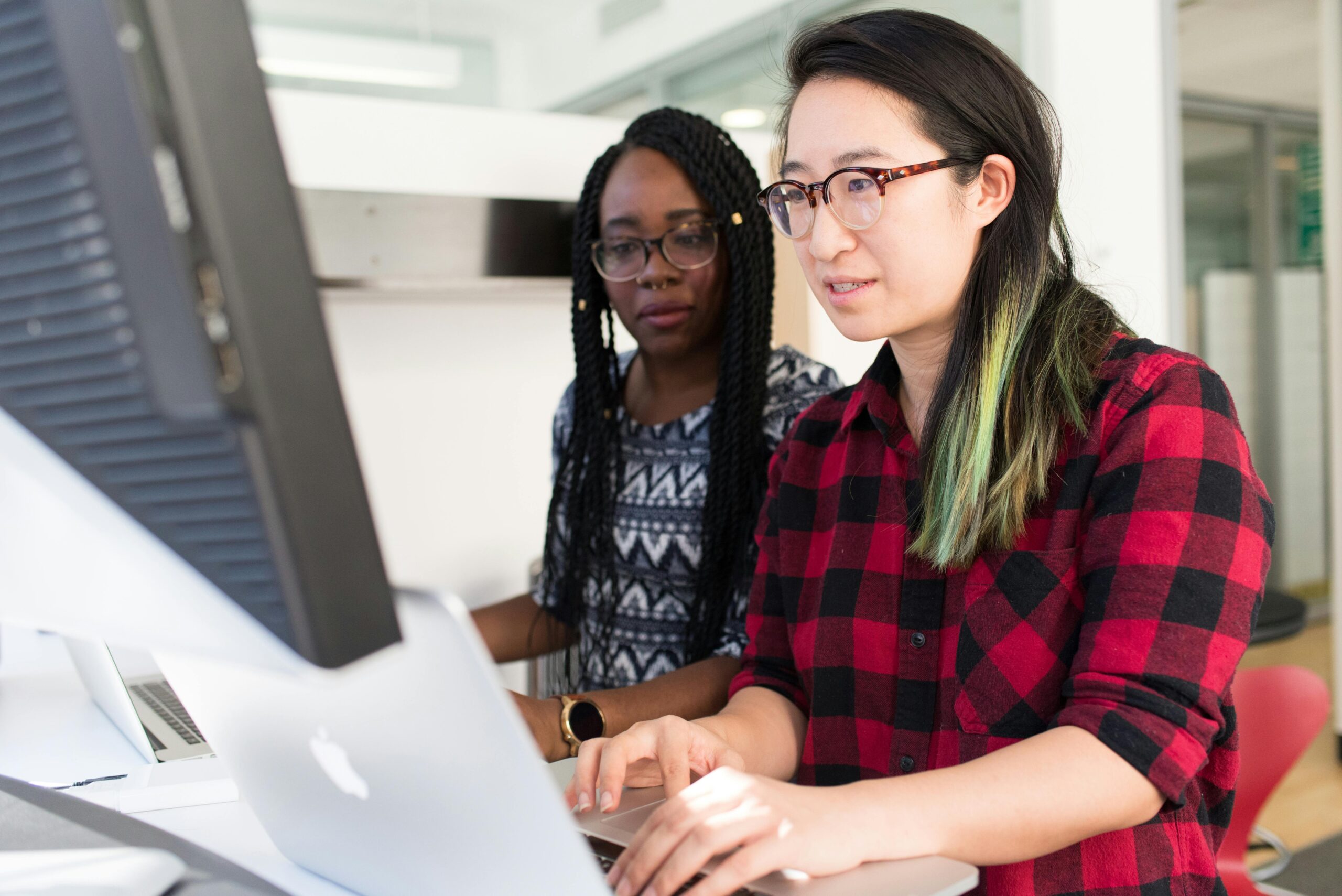 Two women working together at a computer in a modern office, collaborating on a tech project.